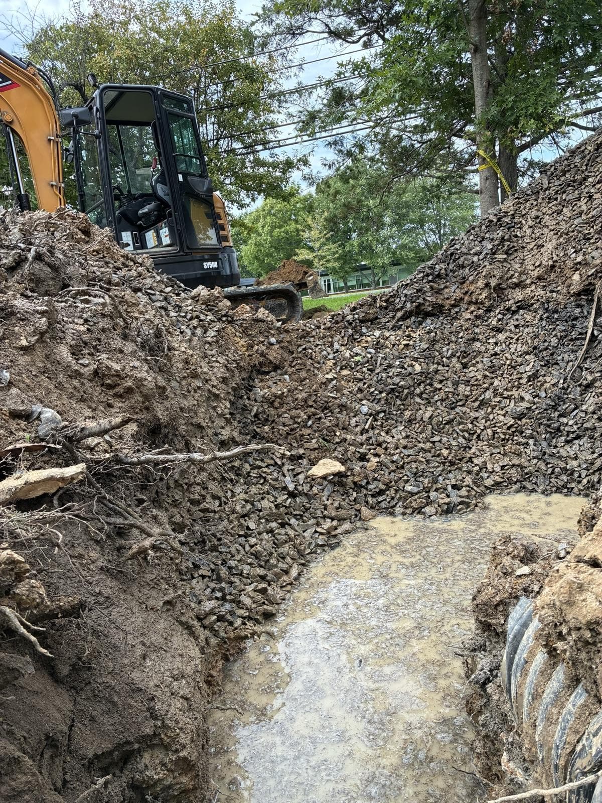 Clean drainage stone backfilled beside a wet drainage trench, with the excavator behind it.