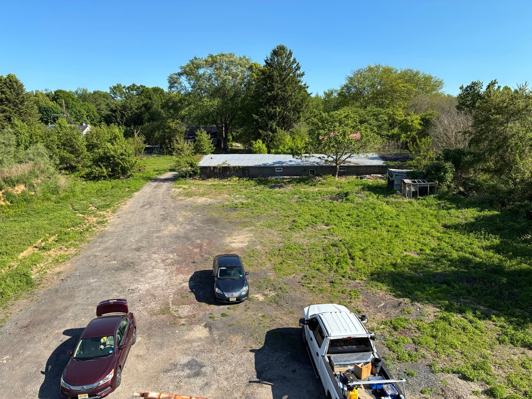 An elevated view of a rough, overgrown lot staged for clearing, with a gravel access drive and surrounding tree line.