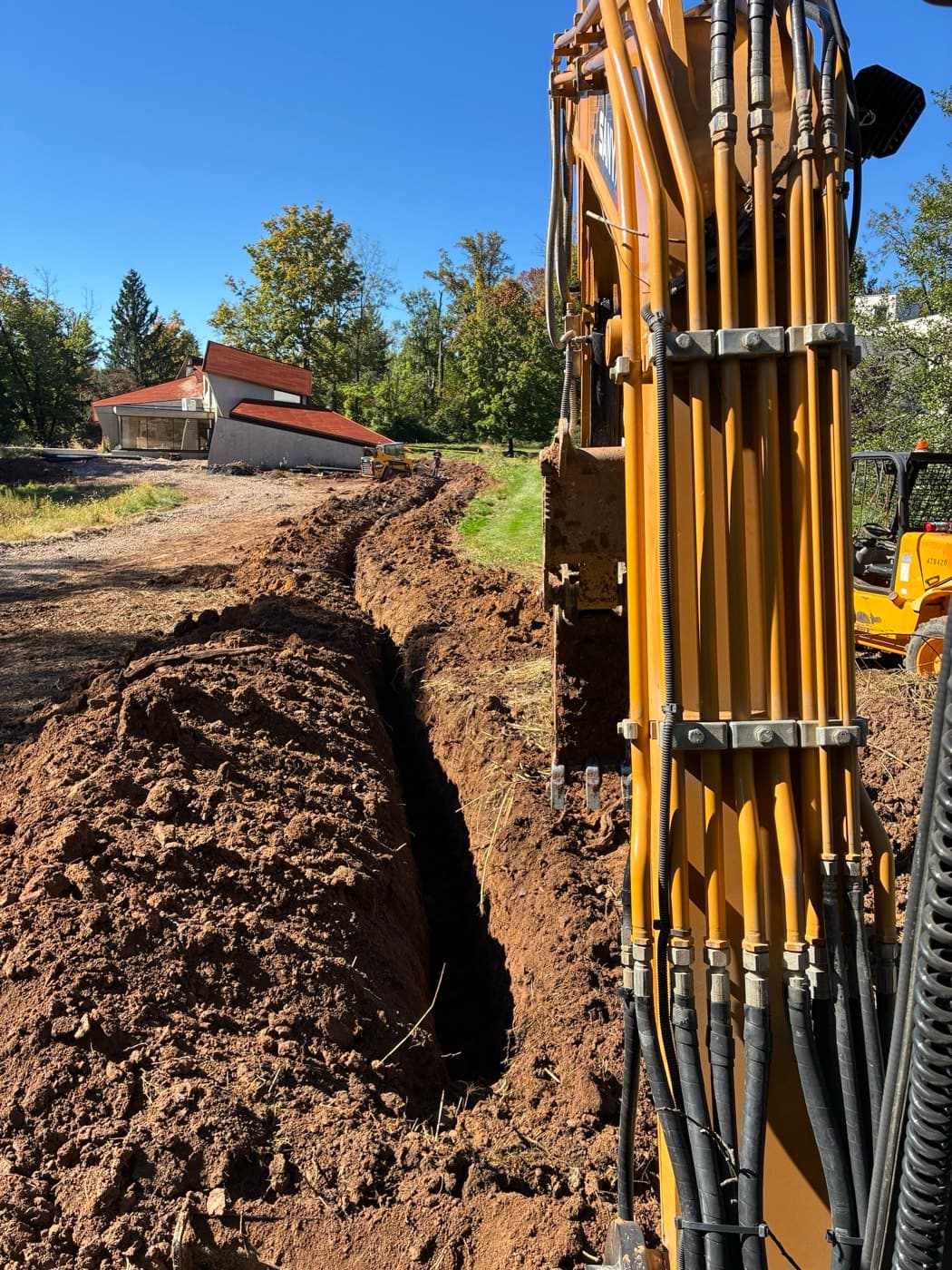A long, single-line utility trench running across a lawn toward the house, cut with an excavator, spoil piled to one side.