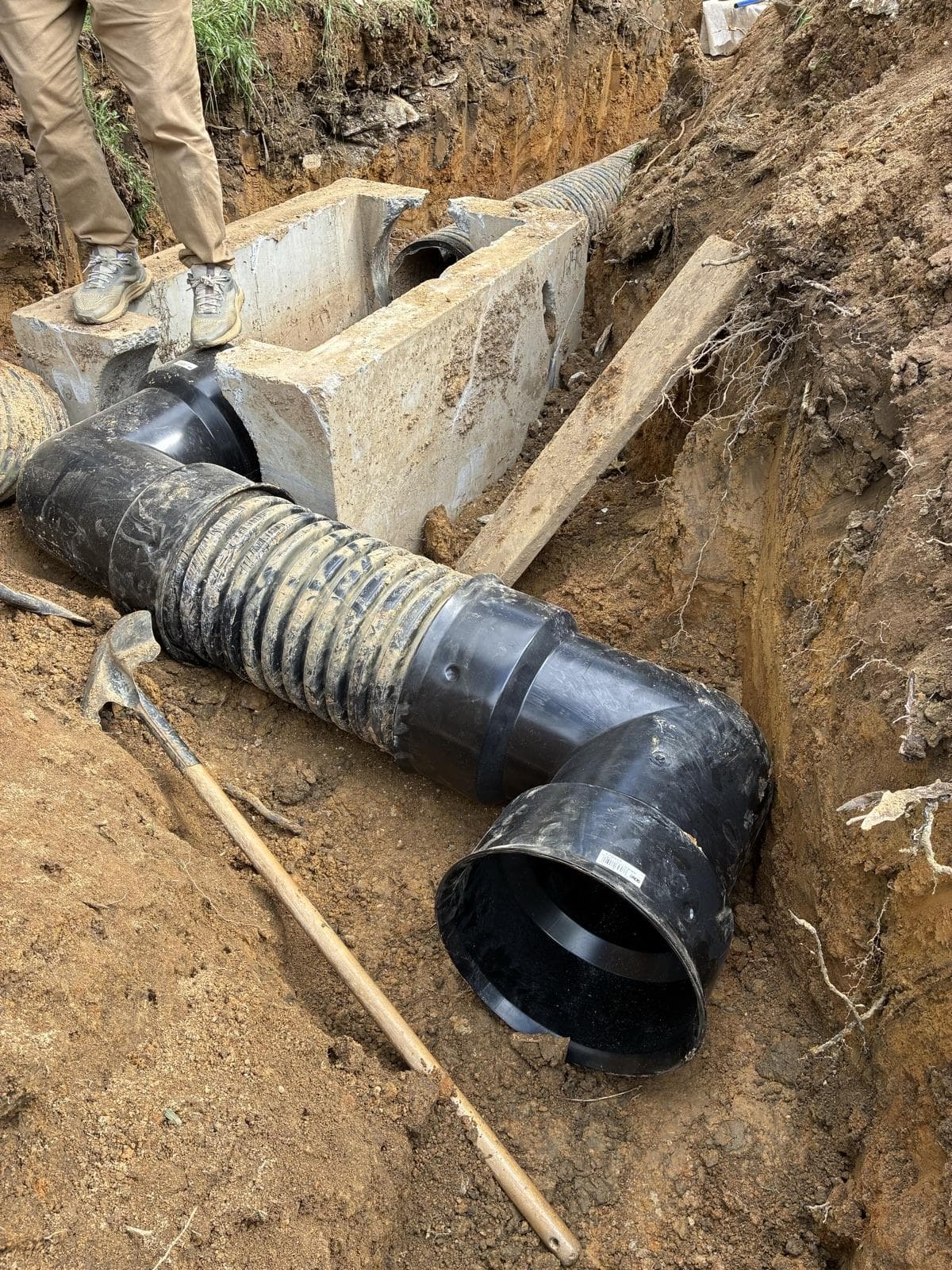 A precast concrete catch basin set in the trench, with corrugated drainage pipe and fittings connected to it.