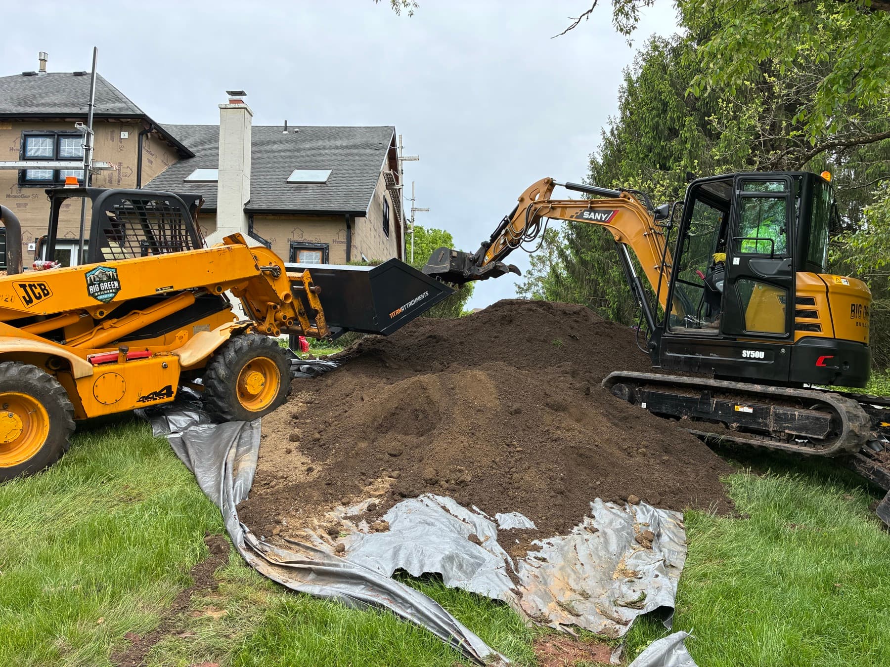 A SANY mini excavator and JCB skid steer working a residential excavation site in the Princeton area, with a fresh soil pile and ground protection over the lawn.