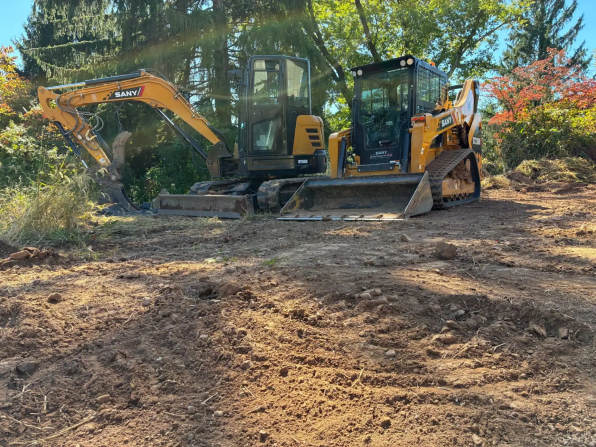 A yellow SANY mini excavator and a SANY compact track loader parked on a freshly cleared lot in the Princeton area, with trees behind.