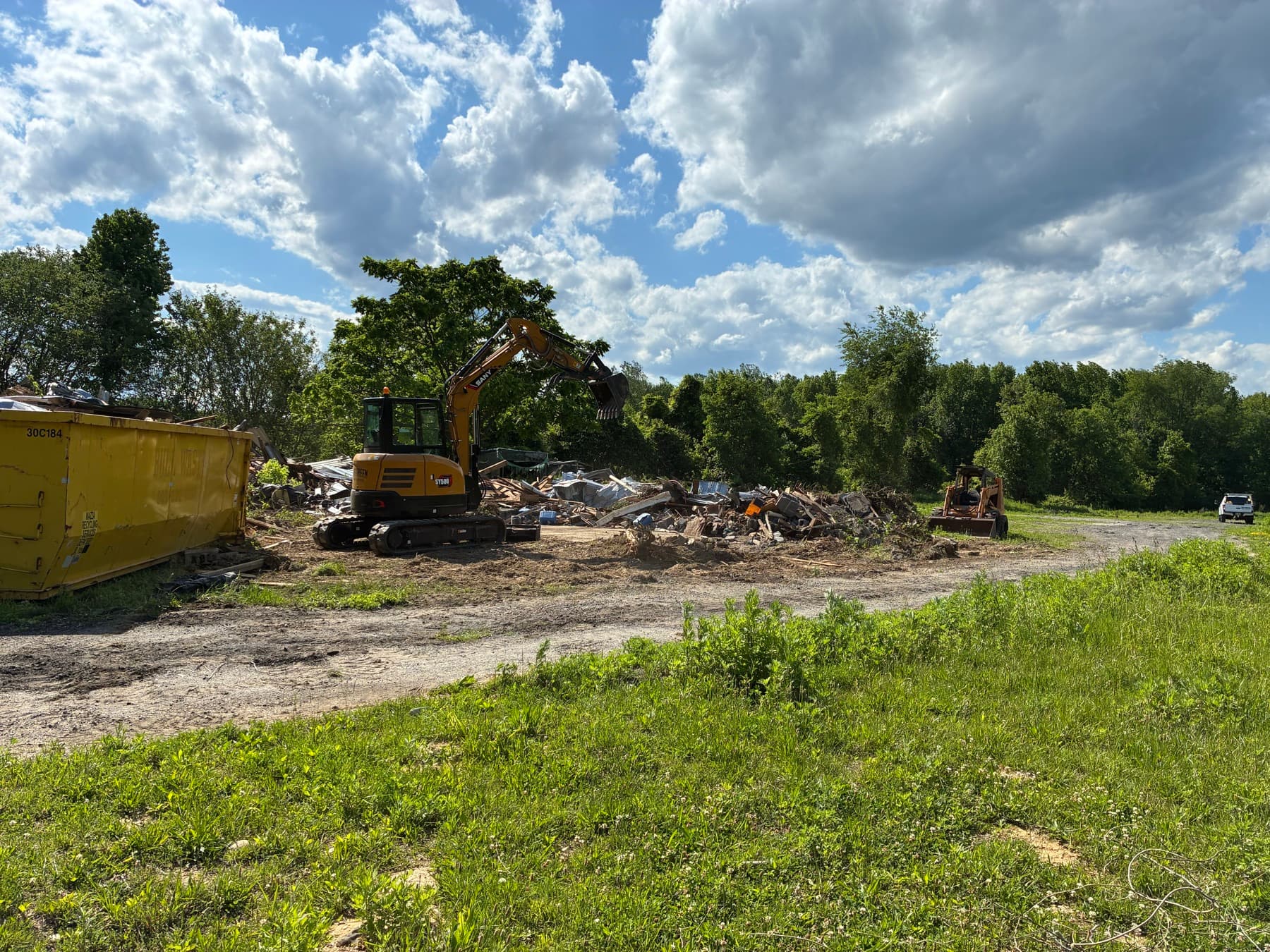 A mini excavator clearing a lot and loading demolition debris into a roll-off dumpster under a bright sky.