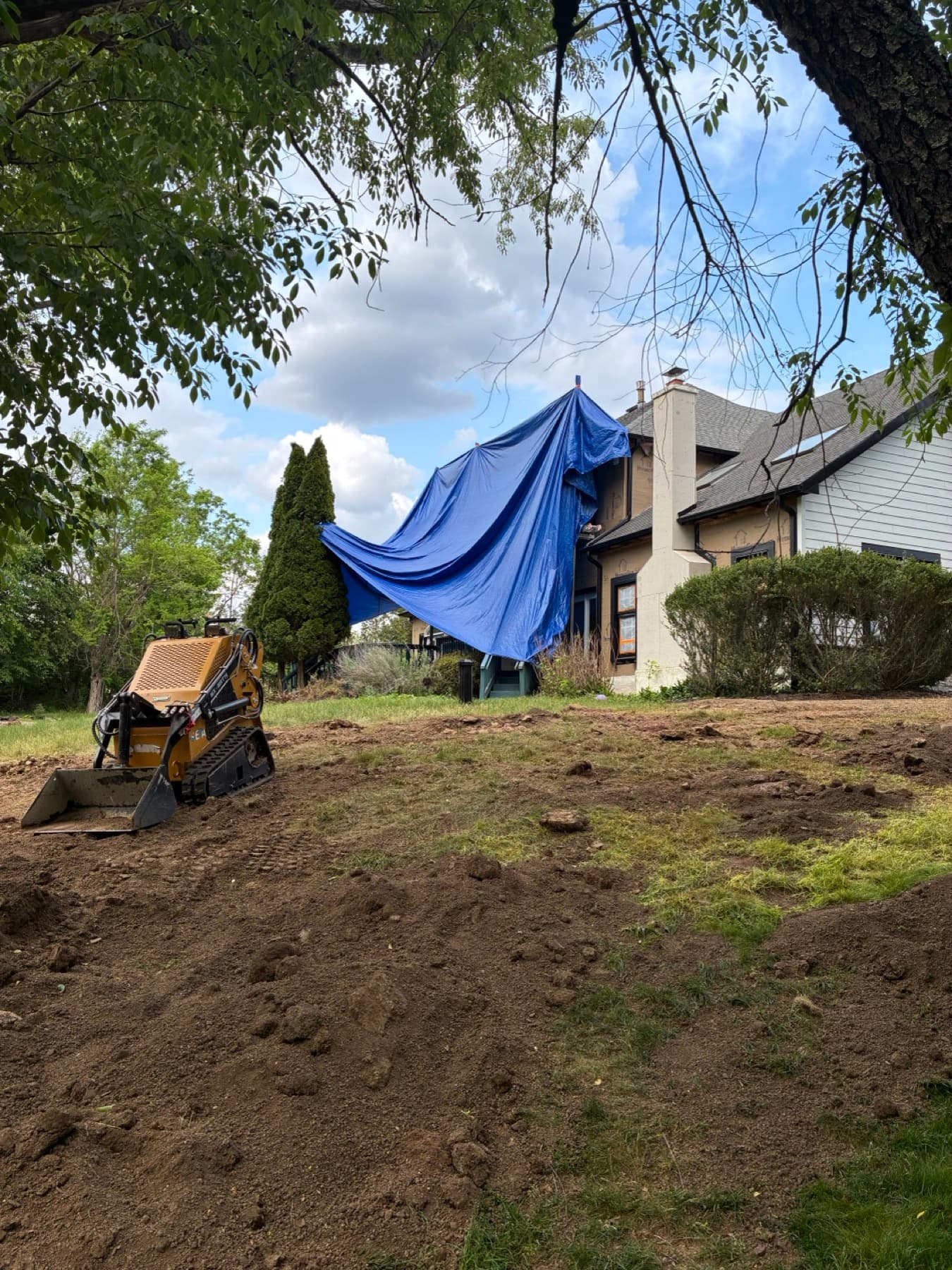 A compact mini skid steer grading bare soil in a residential backyard next to the house, with a protective tarp staged nearby.