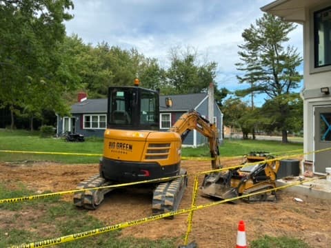 A compact excavator and tracked skid steer working inside a taped-off side yard next to a house.