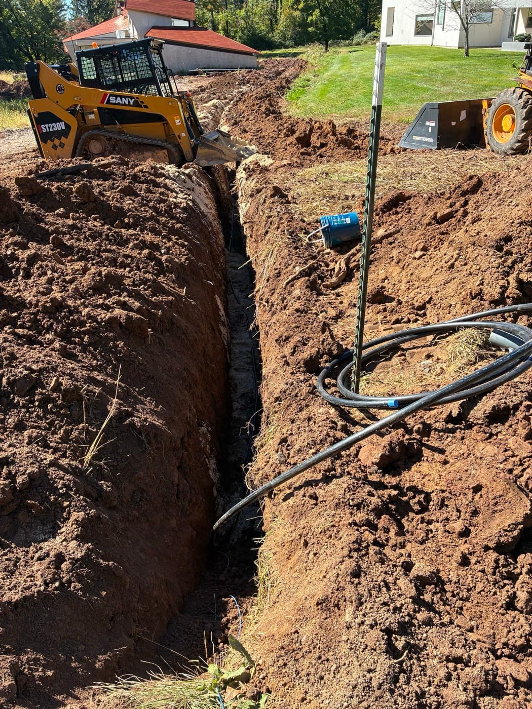 A deep, narrow trench for a new water service line, with the coiled poly pipe and tracer wire staged beside it and a compact track loader at the far end.